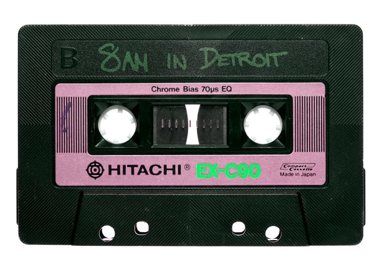 A black cassette tape with a pink label, featuring the text "8AM IN DETROIT" handwritten in green. The brand "HITACHI" and model "EX-90" are printed on the label.