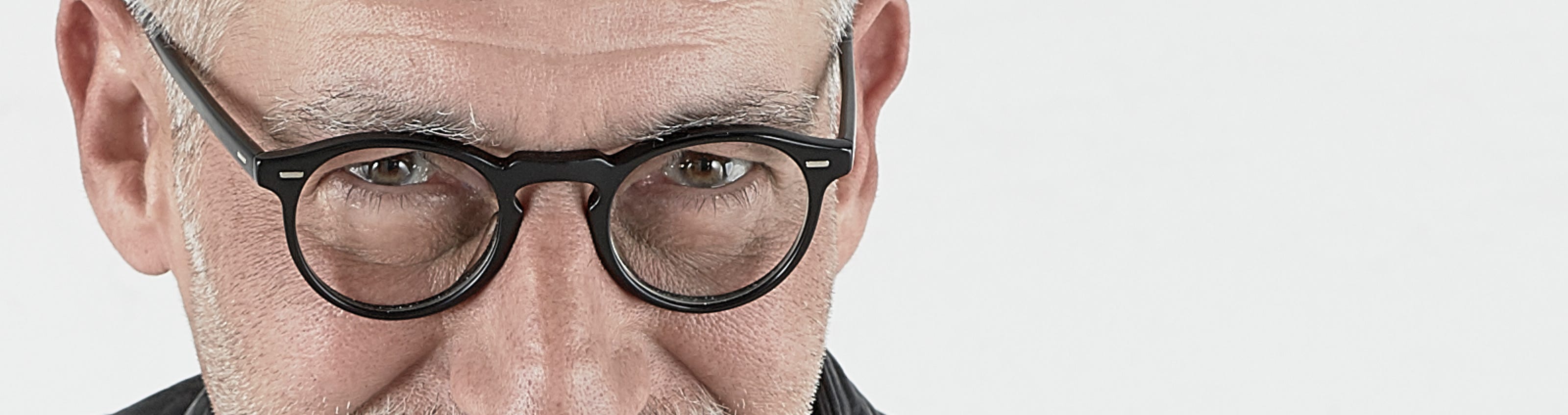A man with glasses, a bald head, and a white beard, wearing a black leather jacket and a white shirt, against a plain background.